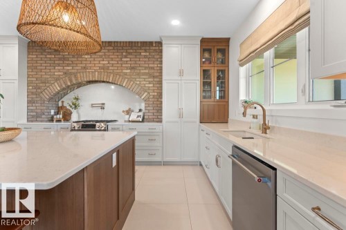 Gourmet kitchen featuring a large central island with light-toned countertop, a brick-faced archway above the range, white cabinetry, a built-in wooden display cabinet, and a stainless steel dishwasher - 2503 Lockhart Way, Cold Lake, AB - Indoor Photo Showing Kitchen