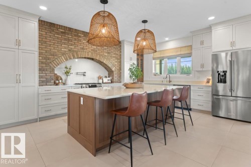 Spacious kitchen featuring a central island with integrated seating, white cabinetry, a large stainless steel refrigerator, light-toned flooring, and a brick-accented wall with an arched cooktop alcove - 2503 Lockhart Way, Cold Lake, AB - Indoor Photo Showing Other Room