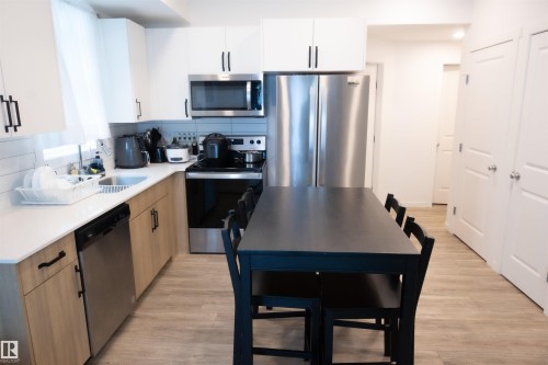 Contemporary kitchen featuring light wood-finish lower cabinetry, white upper cabinetry, stainless steel appliances, and wood-finish flooring - #103 860 Secord Bv Nw, Edmonton, AB - Indoor Photo Showing Kitchen