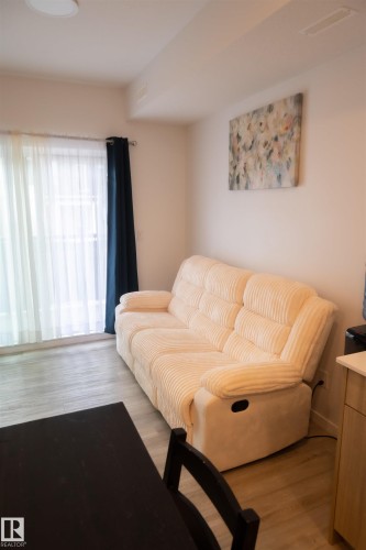 Living area featuring wood-finish flooring, a wall-mounted art piece, and a large window with sheer and blackout curtains - #103 860 Secord Bv Nw, Edmonton, AB - Indoor