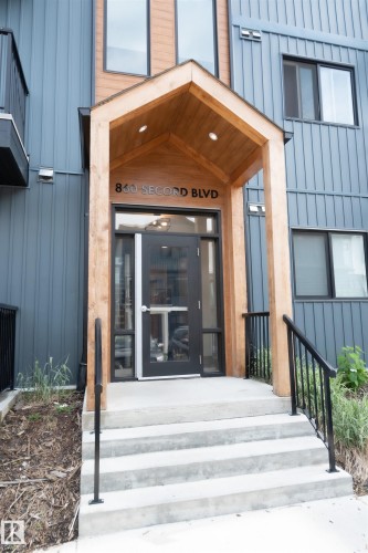 Modern building entrance featuring a wood-clad portico with recessed lighting, concrete steps, and black metal handrails - #103 860 Secord Bv Nw, Edmonton, AB - Outdoor