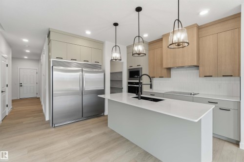 Modern kitchen featuring a large central island with a built-in sink, stainless steel appliances, light wood-finish flooring, two-tone cabinetry, and three pendant light fixtures - 5872 Raven Crescent Nw, Edmonton, AB - Indoor Photo Showing Kitchen With Upgraded Kitchen