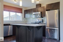 Kitchen featuring a central island with a light-toned countertop, wood-finish flooring, dark wood-finish cabinetry, stainless steel appliances, and a tiled backsplash - 