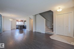 Open-concept living area with dark wood-finish flooring, featuring a kitchen island with pendant lighting, a carpeted staircase with a dark handrail, and a tiled entryway with double bifold doors - 