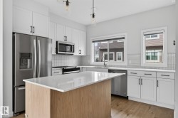 Modern kitchen featuring a light wood-finish island with a white countertop, white cabinetry with black hardware, stainless steel appliances, and a white tile backsplash - 