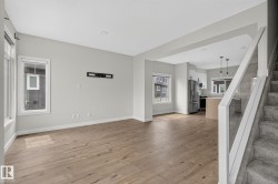 Open-concept living space featuring wood-finish flooring, light grey wall paint, multiple windows with white trim, and a contemporary kitchen with stainless steel appliances - 