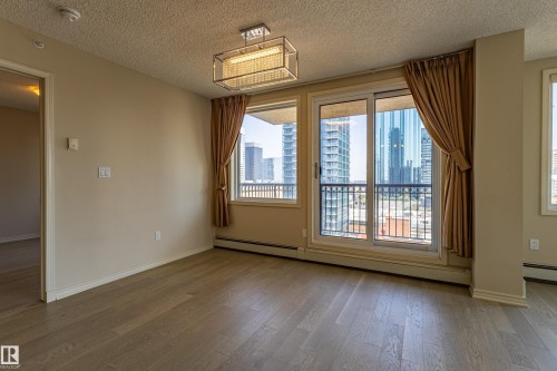 Spacious room featuring wood-finish flooring, neutral paint, a modern ceiling light fixture, and a sliding glass door opening to a balcony with city buildings in the background - #1501 10180 104 St Nw, Edmonton, AB - Indoor Photo Showing Other Room