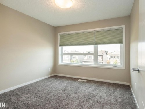 Neutral-toned room featuring a large window with horizontal blinds, textured gray carpet, and white trim - 17263 9 Avenue Sw, Edmonton, AB 