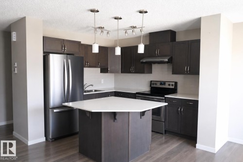 Modern kitchen featuring dark wood-finish cabinetry, stainless steel appliances, a white subway tile backsplash, and a hexagonal island with a light-colored countertop - 17263 9 Avenue Sw, Edmonton, AB 