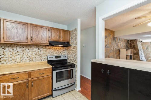 Kitchen featuring wood cabinetry, a mosaic tile backsplash, a stainless steel range, and a pass-through counter with dark wood-finish base cabinets - #112 2904 139 Av Nw, Edmonton, AB - Indoor Photo Showing Kitchen
