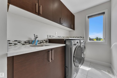Dedicated laundry space featuring dark wood-finish cabinetry, a light-toned countertop with an undermount sink, and a mosaic tile backsplash - 3001 55 Street, Beaumont, AB - Indoor Photo Showing Laundry Room