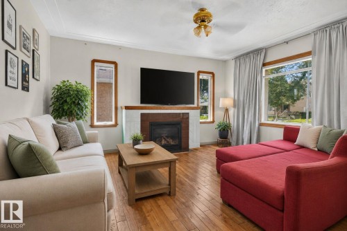 Living area featuring wood-finish flooring, a fireplace with a tiled surround, a large window, a ceiling fan, and crown molding - 12229 88 Street Nw, Edmonton, AB - Indoor Photo Showing Living Room With Fireplace