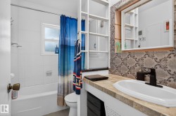 Bathroom featuring a white porcelain tub, white tiled shower surround, frosted window, and a vanity with a white basin sink, tiled countertop, and black faucet - 