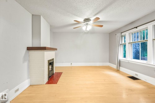 Living area featuring a white brick fireplace with a wood mantle, wood-finish flooring, a ceiling fan, and large multi-pane windows - 9824 91 Avenue Nw, Edmonton, AB - Indoor Photo Showing Other Room With Fireplace
