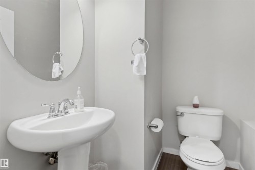 Pedestal sink with chrome faucet, oval mirror, white toilet, and wood-finish flooring - 1528 35 Avenue Nw, Edmonton, AB - Indoor Photo Showing Bathroom
