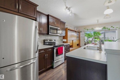 Kitchen featuring dark wood cabinetry, stainless steel appliances, a tile backsplash, light-toned countertops, and wood-finish flooring - 1528 35 Avenue Nw, Edmonton, AB - Indoor Photo Showing Kitchen With Double Sink