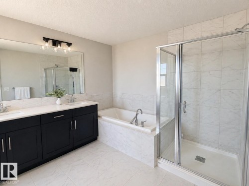 Bathroom featuring a dual vanity with dark cabinetry and white countertops, a built-in soaking tub, a glass-enclosed shower with tiled walls, and light-toned floor tiling - 1327 20 Nw, Edmonton, AB - Indoor Photo Showing Bathroom