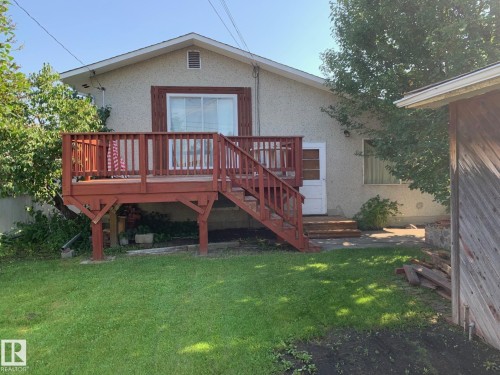 Rear exterior featuring an elevated wood deck with railing and stairs, a stucco finish facade, white-framed windows, and mature trees - 16321 - 109A Ave Nw, Edmonton, AB - Outdoor With Deck Patio Veranda
