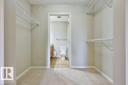 Carpeted closet with white wire shelving, providing access to a bathroom with wood-finish flooring, a vanity with wood cabinetry, a light-toned countertop, and a white toilet - #406 4903 47 Av, Stony Plain, AB - Indoor With Storage