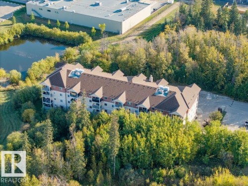 Multi-story building surrounded by mature trees, featuring a brown shingle roof, multiple balconies, and an adjacent parking area - #406 4903 47 Av, Stony Plain, AB - Outdoor With View