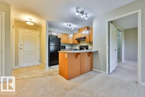 Full kitchen featuring light wood-finish cabinetry, black appliances, track lighting, and a peninsula with countertop seating - #406 4903 47 Av, Stony Plain, AB - Indoor Photo Showing Kitchen