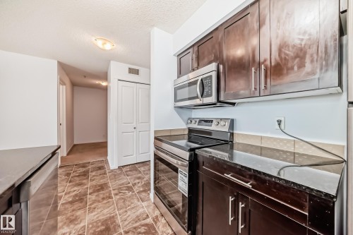 Kitchen featuring dark wood-finish cabinetry, black countertops, and stainless steel appliances including an oven range and microwave - 103 3315 James Mowatt Trail Sw, Edmonton, AB - Indoor Photo Showing Kitchen