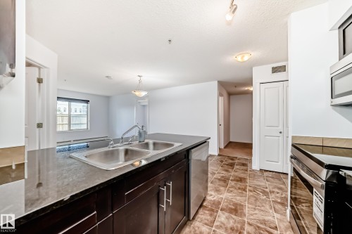 Kitchen featuring a double basin stainless steel sink, dark cabinetry, and stone countertops - 103 3315 James Mowatt Trail Sw, Edmonton, AB - Indoor Photo Showing Kitchen With Double Sink