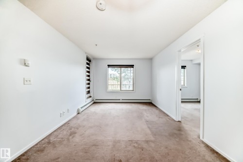 Spacious room featuring neutral carpeting, white painted walls, and a large window with horizontal blinds - 103 3315 James Mowatt Trail Sw, Edmonton, AB - Indoor Photo Showing Other Room