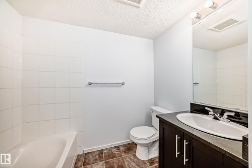 Full bathroom featuring a white porcelain tub with tiled surround, dark wood-finish vanity with an integrated sink, polished chrome faucet, and a large wall-mounted mirror - 103 3315 James Mowatt Trail Sw, Edmonton, AB - Indoor Photo Showing Bathroom