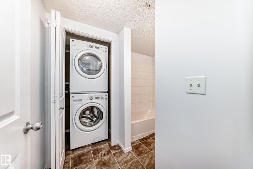 Integrated stackable washer and dryer unit, complemented by brown-toned tile flooring and a white tiled shower surround with a bathtub - 103 3315 James Mowatt Trail Sw, Edmonton, AB - Indoor Photo Showing Laundry Room