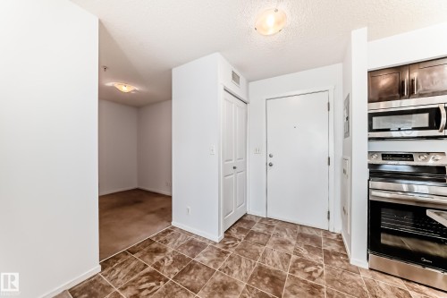 Entry area featuring square tile flooring, a white entry door, a bi-fold closet door, and a kitchen wall with dark wood-finish cabinetry and stainless steel appliances - 103 3315 James Mowatt Trail Sw, Edmonton, AB - Indoor