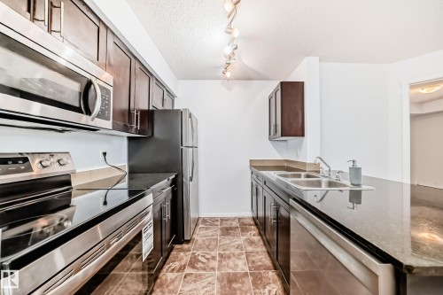 Galley kitchen featuring stainless steel appliances, dark wood-finish cabinetry, stone-finish countertops, and ceramic tile flooring - 103 3315 James Mowatt Trail Sw, Edmonton, AB - Indoor Photo Showing Kitchen With Double Sink
