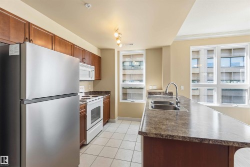 Kitchen featuring abundant wood-finish cabinetry, stainless steel refrigerator, white electric range with matching microwave, dual basin sink, and tile flooring - #708 9939 109 St Nw, Edmonton, AB - Indoor Photo Showing Kitchen With Double Sink
