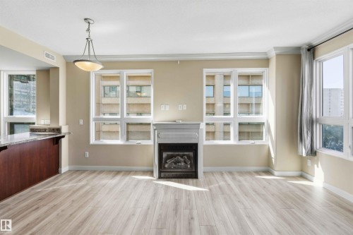 Spacious living area featuring light wood-finish flooring, a contemporary fireplace with mantel, and multiple large windows - #708 9939 109 St Nw, Edmonton, AB - Indoor With Fireplace