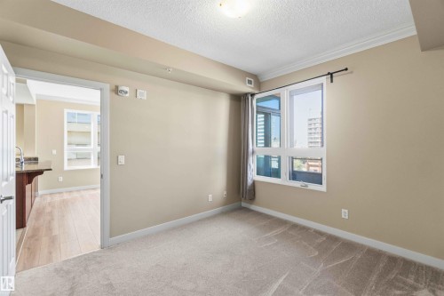 Carpeted room featuring a large window, crown molding, and a doorway opening to a space with wood-finish flooring and a kitchen counter with an undermount sink - #708 9939 109 St Nw, Edmonton, AB - Indoor Photo Showing Other Room