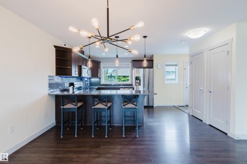 Open-concept kitchen featuring an island with a breakfast bar, dark wood-finish cabinetry, stainless steel appliances, a tile backsplash, and wood-finish flooring - 6717 Elston Lane Nw, Edmonton, AB - Indoor