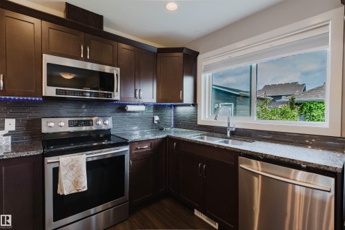 Kitchen featuring dark wood cabinetry, stainless steel appliances, speckled stone countertops, a tile backsplash, and a large window - 6717 Elston Lane Nw, Edmonton, AB - Indoor Photo Showing Kitchen With Stainless Steel Kitchen With Double Sink