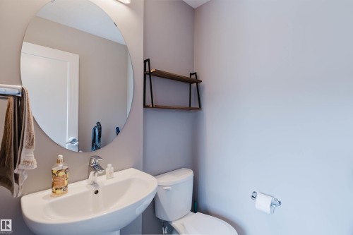 Compact bathroom featuring a pedestal sink with chrome faucet, circular frameless mirror, wall-mounted light fixture, and built-in shelving with wood-finish surfaces and black metal supports - 6717 Elston Lane Nw, Edmonton, AB - Indoor Photo Showing Bathroom