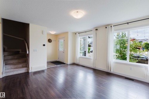 Spacious entry and living area featuring dark wood-finish flooring, multiple windows with white trim, a front door with glass inserts, a carpeted staircase with white railings, and an overhead flush-mount light fixture - 6717 Elston Lane Nw, Edmonton, AB - Indoor
