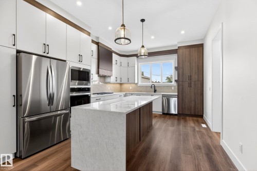 Modern kitchen featuring a large waterfall island with a stone countertop, wood-finish flooring, two-tone cabinetry, and stainless steel appliances including a built-in oven and microwave - 5308 103A Avenue, Edmonton, AB - Indoor Photo Showing Kitchen With Upgraded Kitchen