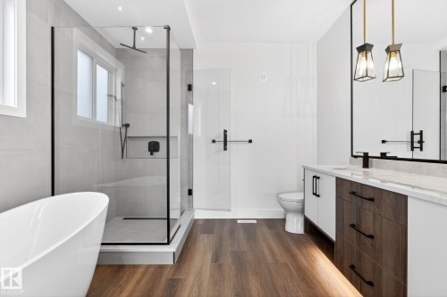 Contemporary bathroom featuring a freestanding soaking tub, a frameless glass shower enclosure with matte black fixtures, wood-finish flooring, a dual-sink vanity with a light-colored countertop, and modern pendant lighting - 5308 103A Avenue, Edmonton, AB - Indoor Photo Showing Bathroom