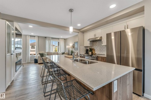 Open-concept living space featuring wood-finish flooring, a kitchen island with seating, stainless steel appliances, a subway tile backsplash, and a built-in fireplace - #25 1530 Tamarack Bv Nw, Edmonton, AB - Indoor Photo Showing Kitchen With Double Sink With Upgraded Kitchen