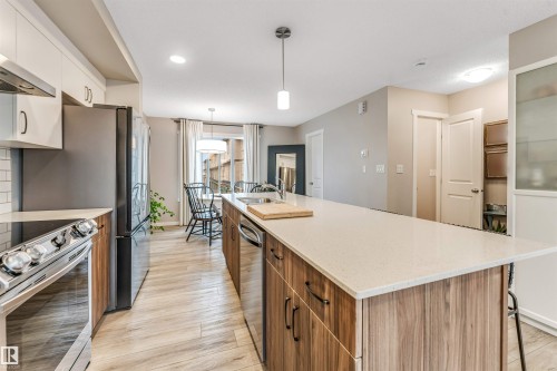 Spacious kitchen featuring a large central island with light-toned countertop, wood-finish cabinetry, stainless steel appliances, and light wood-finish flooring - #25 1530 Tamarack Bv Nw, Edmonton, AB - Indoor Photo Showing Kitchen