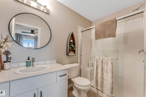 Bathroom featuring a shower with a sliding glass door, a vanity with a white countertop and integrated sink, a circular mirror, and wood-finish flooring - #25 1530 Tamarack Bv Nw, Edmonton, AB - Indoor Photo Showing Bathroom