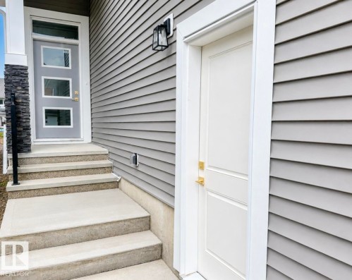 Exterior featuring horizontal siding, a contemporary entry door with three glass panels, a stone-accented pillar, and concrete steps with an integrated black metal handrail - 8423 Mayday Link Link Sw, Edmonton, AB - Outdoor With Exterior