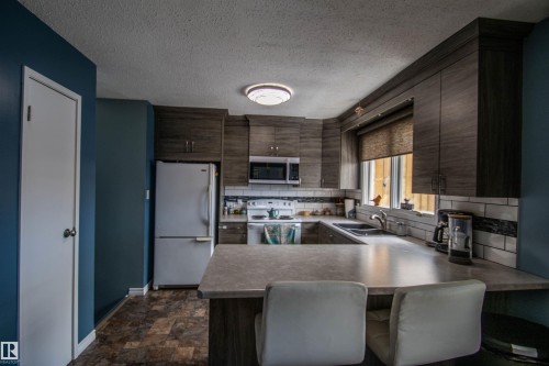 Modern kitchen featuring dark wood-finish cabinetry, an integrated microwave, a double basin sink, and a multi-toned subway tile backsplash - 5017 55 Ave, St. Paul Town, AB - Indoor Photo Showing Kitchen With Double Sink