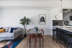 Open-concept living area featuring light wood-finish flooring, a contemporary kitchen with dark cabinetry and a tiled backsplash, a kitchen island with seating, a dining area, and a modern light fixture - 