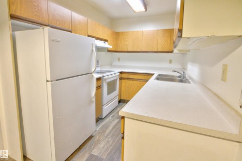 Compact kitchen featuring wood-finish cabinetry, white countertops, a double basin stainless steel sink, and wood-finish flooring - #302 47 Akins Dr, St. Albert, AB - Indoor Photo Showing Kitchen With Double Sink