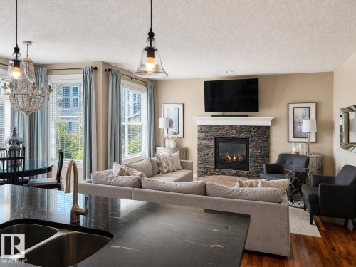 Open-concept living area featuring a stacked stone fireplace with a white mantel, rich wood-finish flooring, and multiple windows with blinds - 2337 Frezenberg Avenue Nw, Edmonton, AB - Indoor Photo Showing Living Room With Fireplace