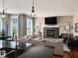 Open-concept living area featuring a stacked stone fireplace with a white mantel, rich wood-finish flooring, and multiple windows with blinds - 